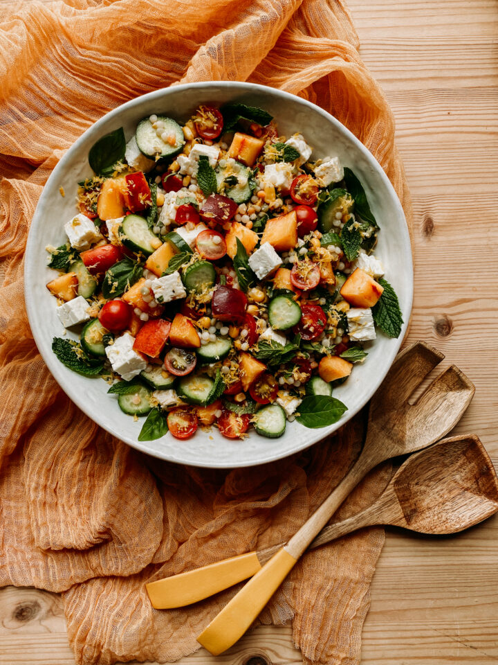 Couscous Summer Salad in a white servinf bowl resting on a yellow cloth with wooden salad servers resting next to the bowl
