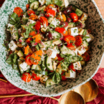 overhead shot of crunchy greek salad with avocado, feta, cucumbers, tomatoes, peppers, olives, and fresh herbs in a green speckled bowl