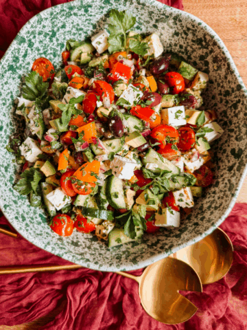 overhead shot of crunchy greek salad with avocado, feta, cucumbers, tomatoes, peppers, olives, and fresh herbs in a green speckled bowl