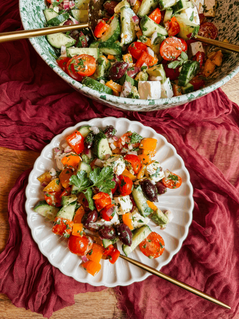 greek salad served in a large green bowl and on a white plate with cucumbers, tomatoes, peppers, olives, avocado, and feta