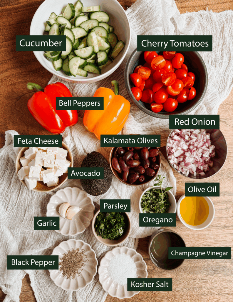 ingredients for greek salad with avocado and feta including cucumbers, cherry tomatoes, bell peppers, feta, olives, avocado, red onion, garlic, herbs, olive oil, vinegar, salt, and pepper
