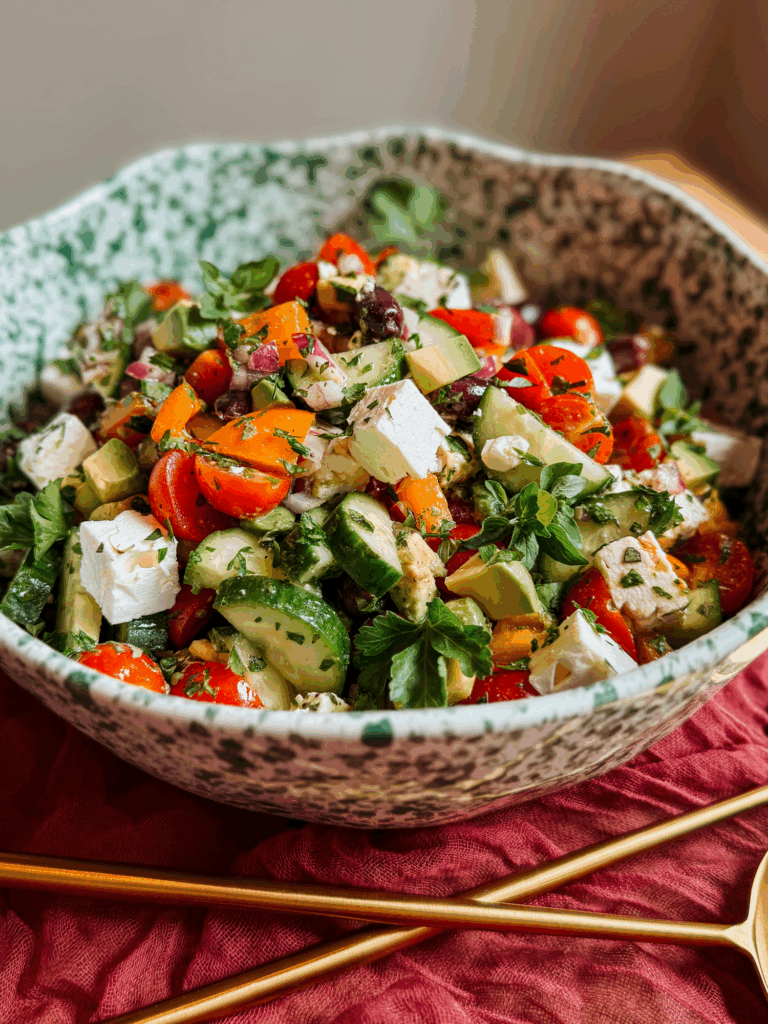 close-up of crunchy greek salad with avocado, feta, olives, cucumbers, tomatoes, and peppers garnished with fresh herbs