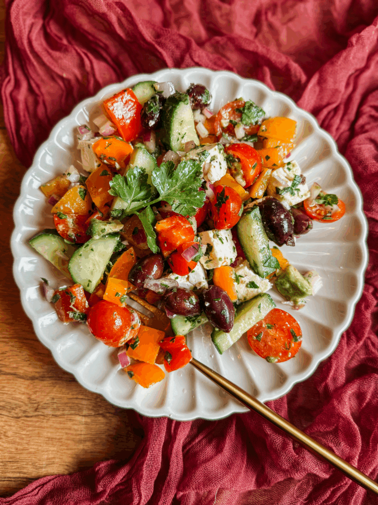 plated greek salad with avocado, feta, cucumbers, tomatoes, peppers, and olives on a white scalloped plate