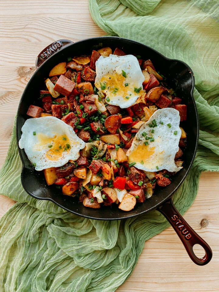 A skillet of Corned Beef Hash topped with three fried eggs resting on a gauzy green cloth