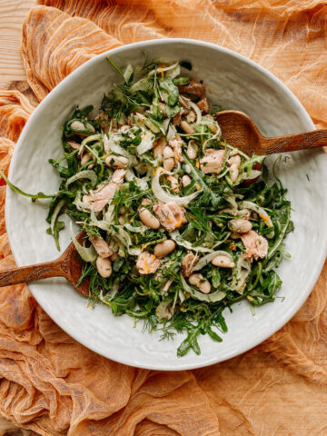 wooden salad utensils resting in a big white salad bowl filled with tuna, arugula, beans, and fennel