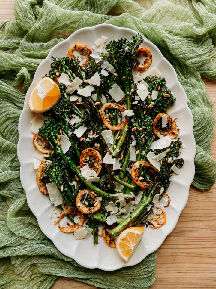 Charred Broccolini with Lemon, Pine Nuts, and Parmesan on a large white platter resting on a gauzy green cloth