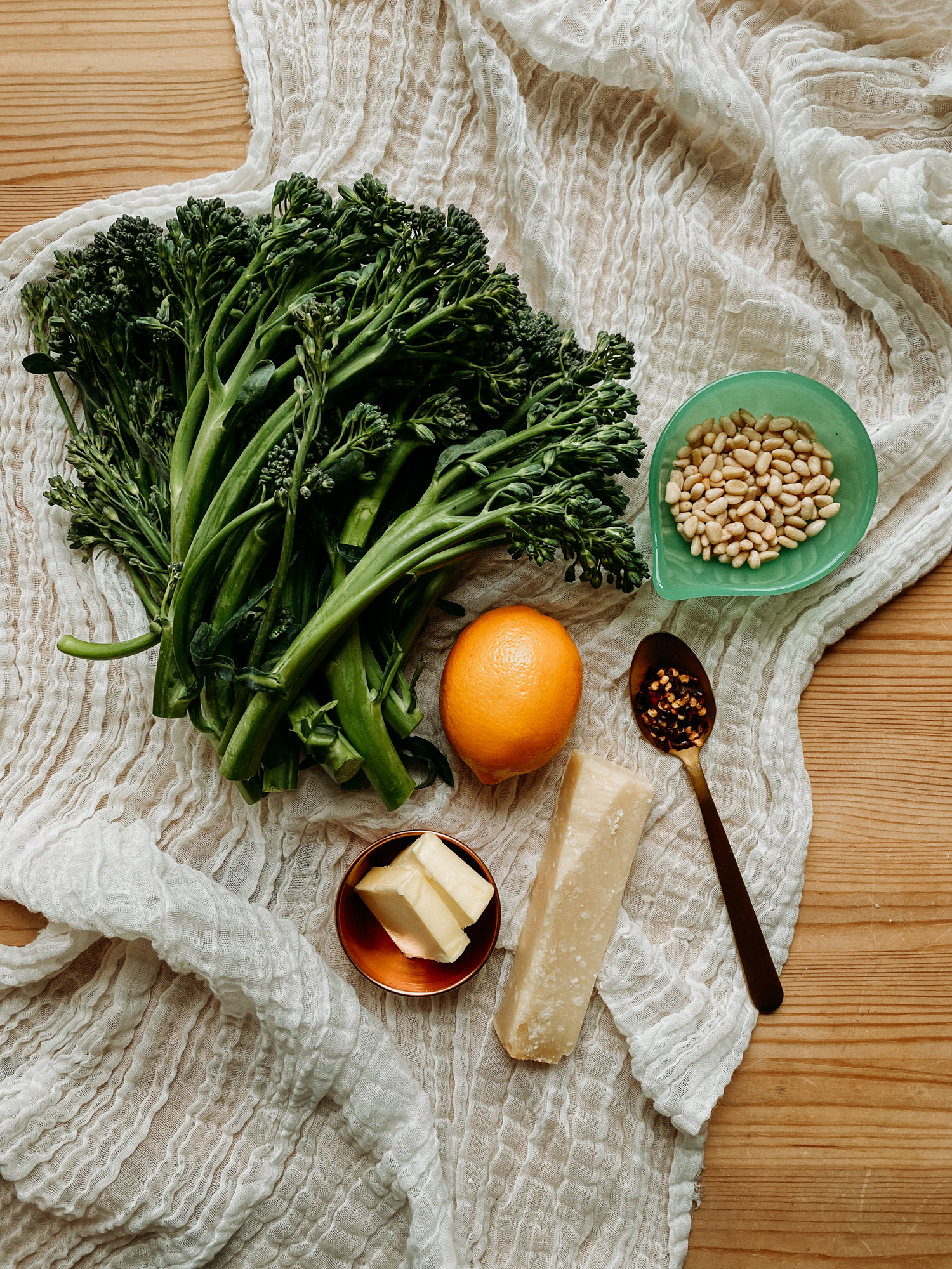 a whit gauzy cloth with raw broccolini, a lemon, pine nuts, a wedge of parmesan, butter, and a spoonful of red pepper flakes