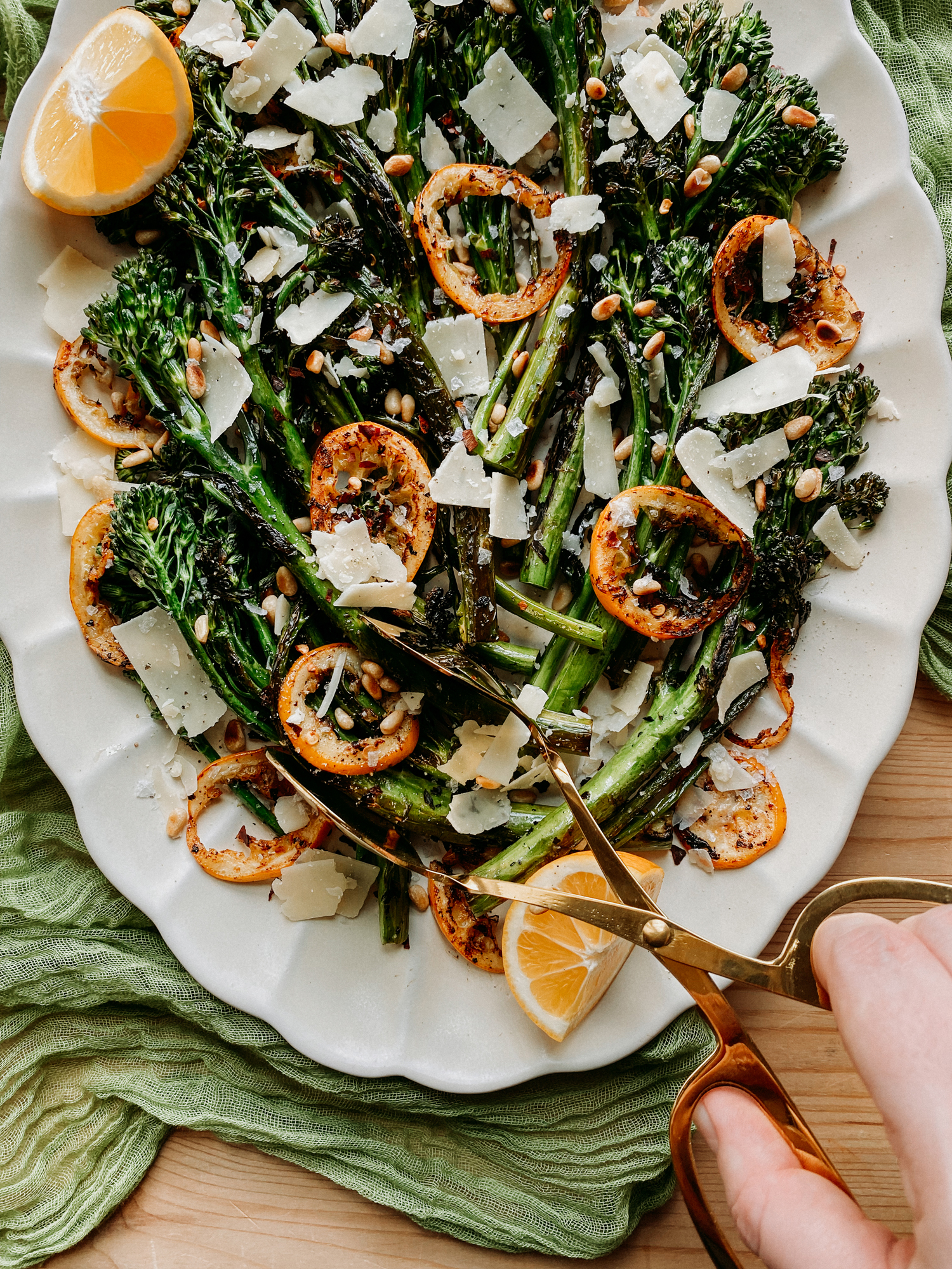 a hand using gold tongs to pick up broccolini off of a white platter