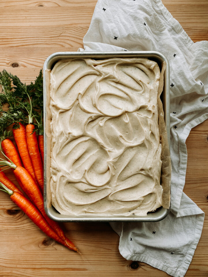 a large Sourdough Discard Carrot Cake with Browned Butter Cream Cheese Frosting restign on a wooden surface with fresh carrots