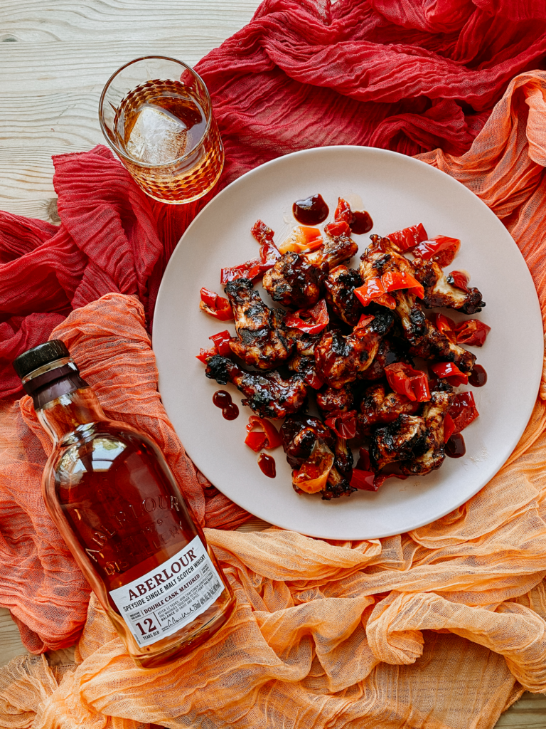 a plate of grilled chicken wings resting next to a bottle of scotch and a glass of whisky on red and orange cloths