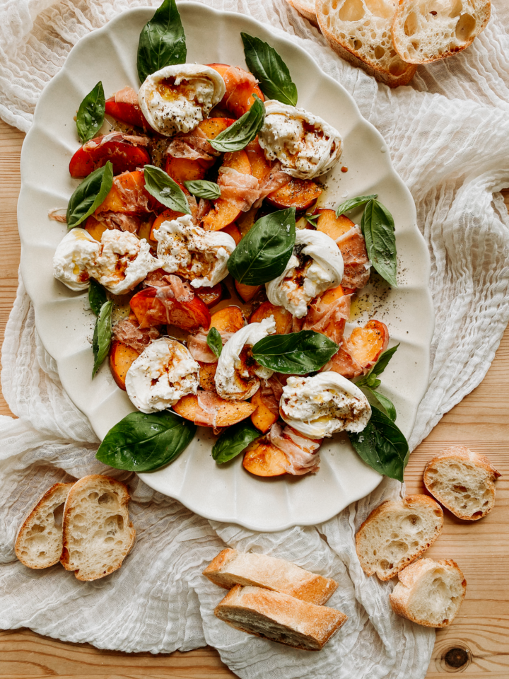 a large white platter with Peach, Prosciutto, and Burrata Salad on top surrounded by sliced baguette