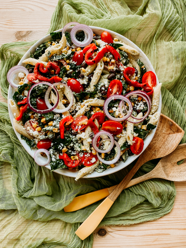 a large white bowl with Southwest Pasta Salad resting on a gauzy green cloth with wooden salad servers
