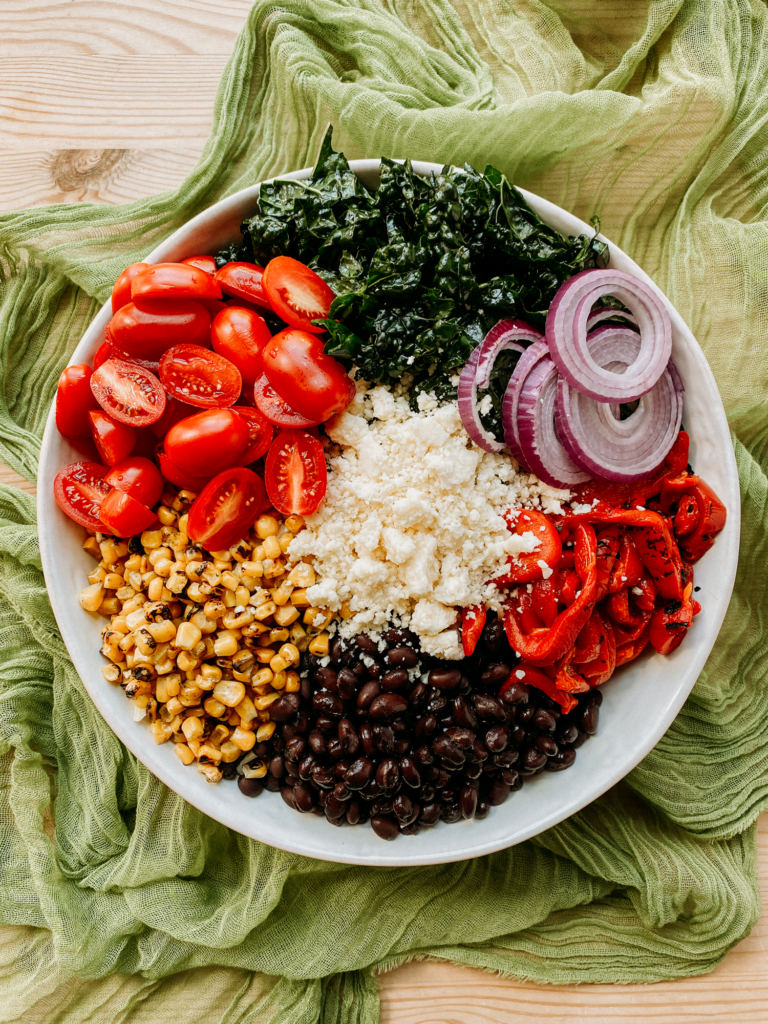 a large white bowl filled with tomatoes, cheese, onions, kale, beans, peppers, and corn