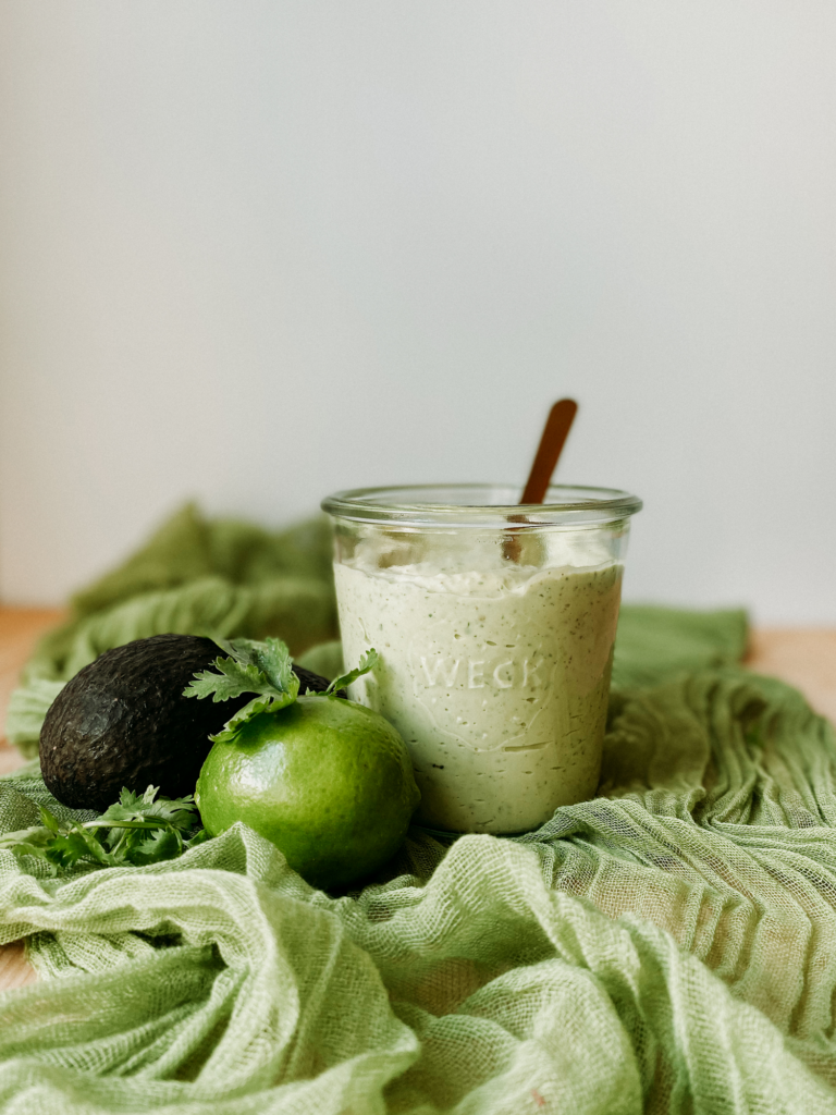 a jar of avocado lime ranch on a gauzy green cloth with an avocado, lime, and cilantro resting nearby