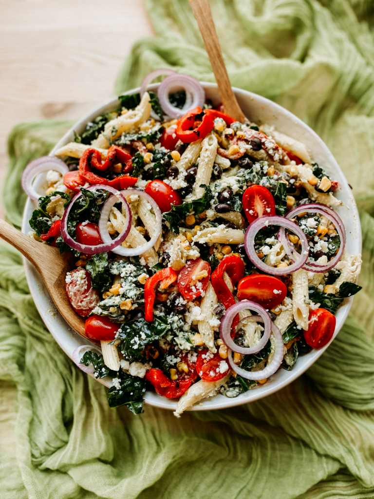wooden utensils resting in a large bowl of pasta salad with veggies and beans on a green cloth