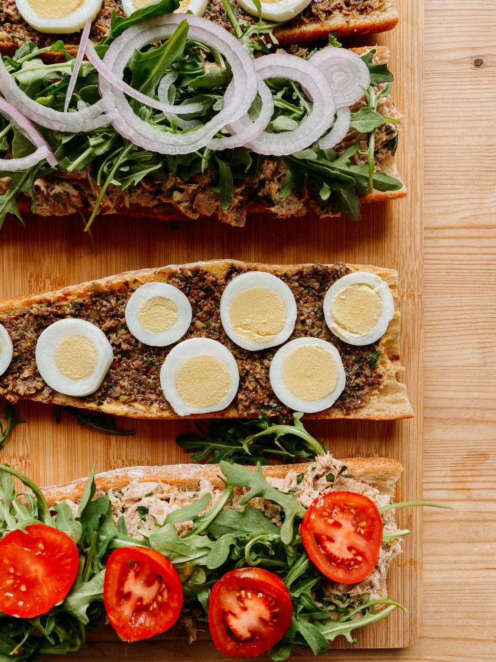 an open baguette with onion, arugula, eggs, tapenade, tuna salad, tomatoes