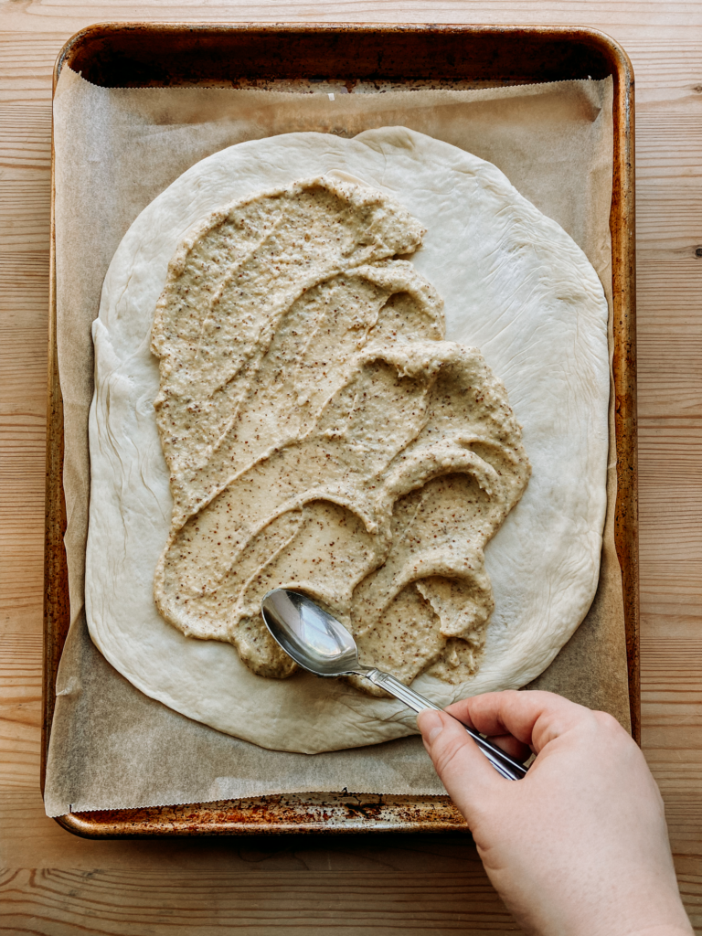 a hand spreading mustard cream sauce over pizza dough with a spoon
