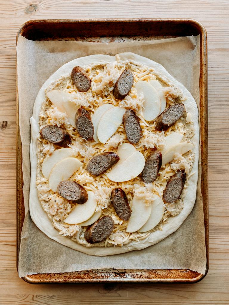 pizza dough topped with gouda cheese, sauerkraut, brat, and apples