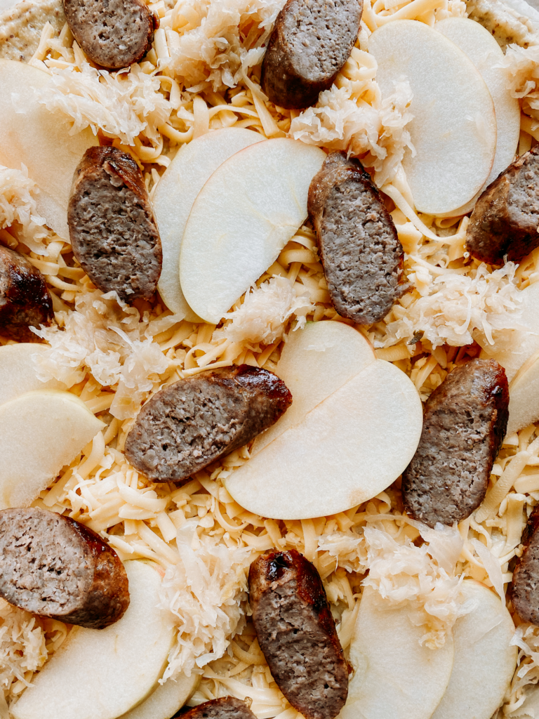 slices of bratwurst and apples on top of a pizza with sauerkraut