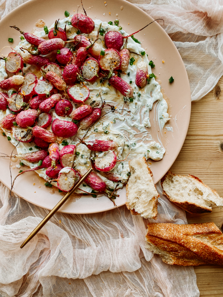 a gold spoon resting on a pink platter with Roasted Radishes with Garlic Chive Yogurt. crusty bread laying nearby on a wood surface.
