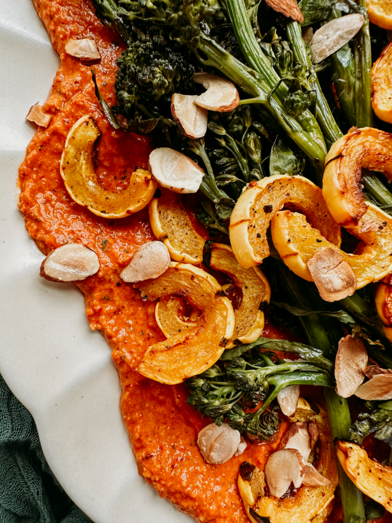 a bed of romesco sauce on a white plate topped with delicata, almonds, and broccolini