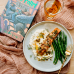 A Harry Potter book resting next to a plate with salmon, mashed potato, and green beans on a pink cloth