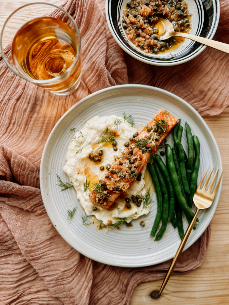a plate with salmon, pommes puree, and green beans next to a glass of white wine on a pink cloth