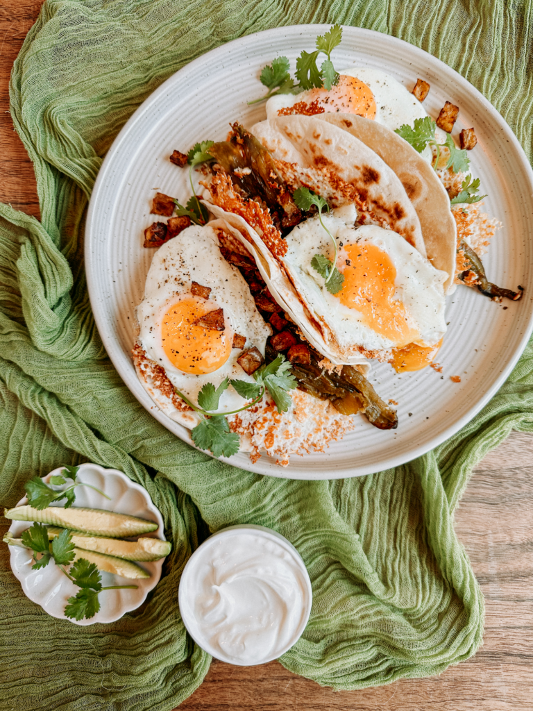 a white plate resting on a green cloth with breakfast tacos on top and avocado and sour cream nearby