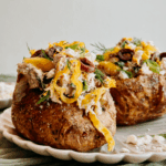 Close-up of Greek tuna salad baked potato topped with feta, olives, pepperoncini, and fresh dill on a scalloped plate.