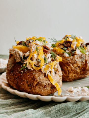 Close-up of Greek tuna salad baked potato topped with feta, olives, pepperoncini, and fresh dill on a scalloped plate.