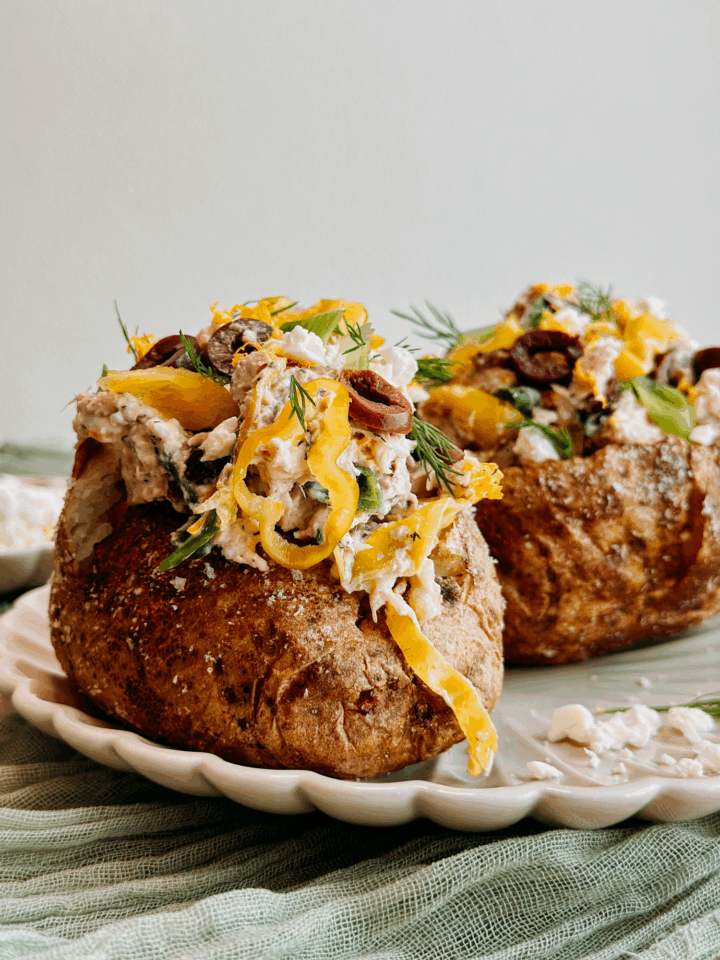 Close-up of Greek tuna salad baked potato topped with feta, olives, pepperoncini, and fresh dill on a scalloped plate.