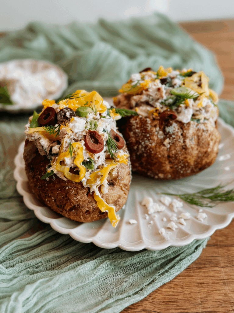 Two salt-crusted baked potatoes filled with Greek tuna salad on a white plate with green cloth backdrop.