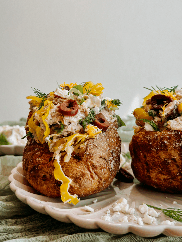 Tall side view of two loaded baked potatoes filled with Greek tuna salad and garnished with pepperoncini, dill, and feta.