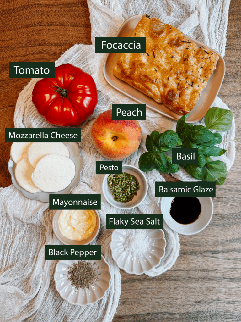 Labeled flat lay of ingredients for peach caprese sandwich including focaccia, tomato, peach, basil, mozzarella, pesto, mayonnaise, and balsamic glaze