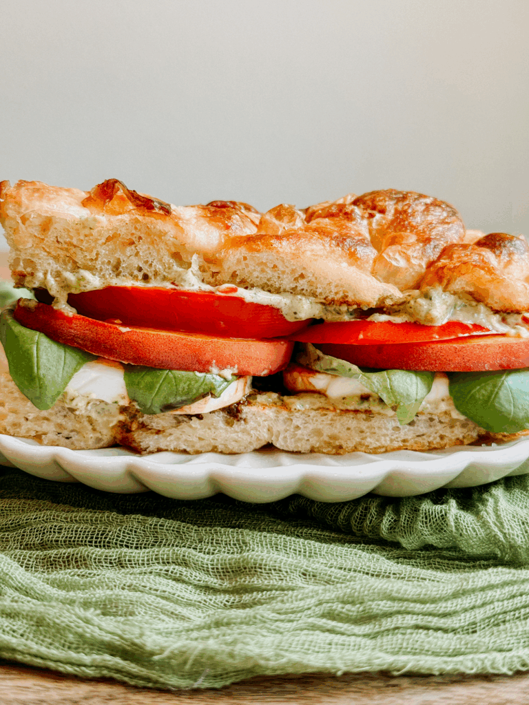 Close-up of peach caprese sandwich with visible layers of tomato, basil, mozzarella, and pesto mayo on toasted focaccia