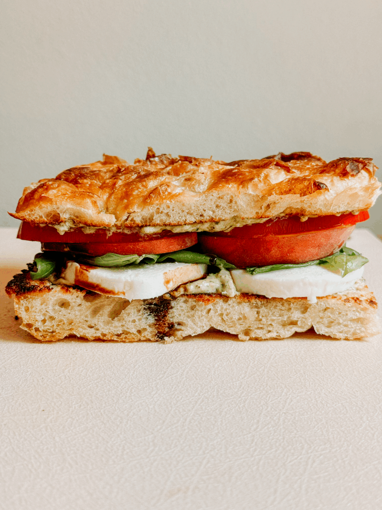Side view of Peach Caprese Sandwich on focaccia, layered with tomato, peach, basil, mozzarella, and pesto mayo.