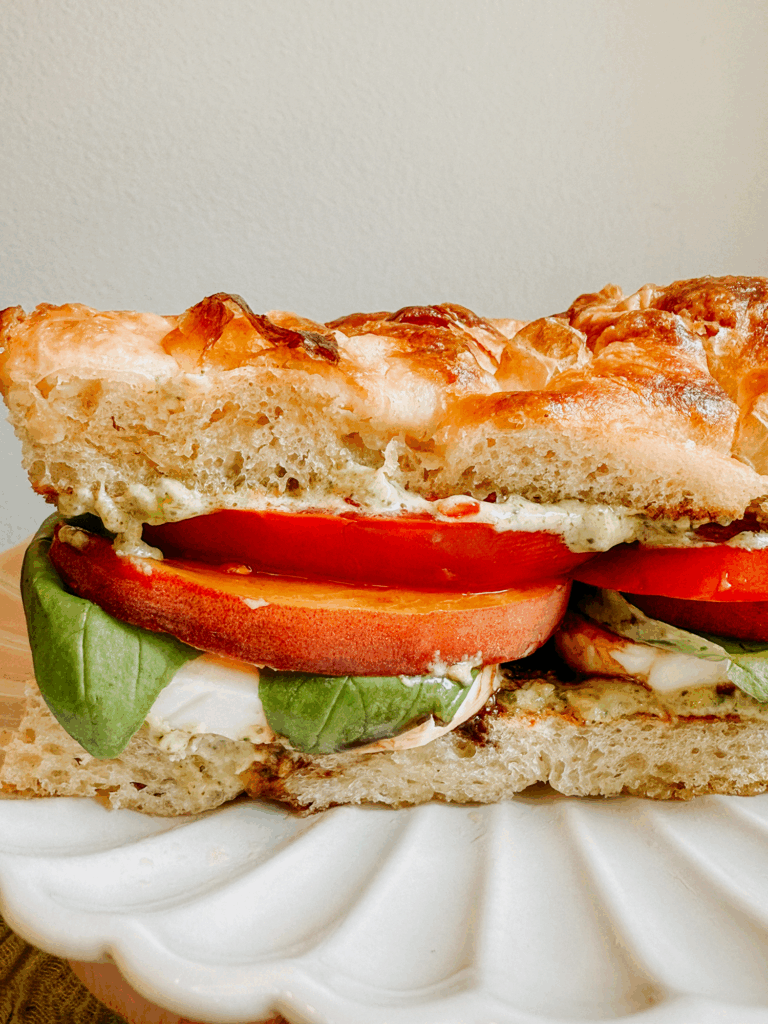 Peach Caprese Sandwich on a scalloped white plate, layered with basil, peach, tomato, and mozzarella on toasted focaccia.
