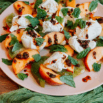 Plate of burrata with peaches, green heirloom tomatoes, chili crisp, and fresh herbs on a light pink plate with green fabric.