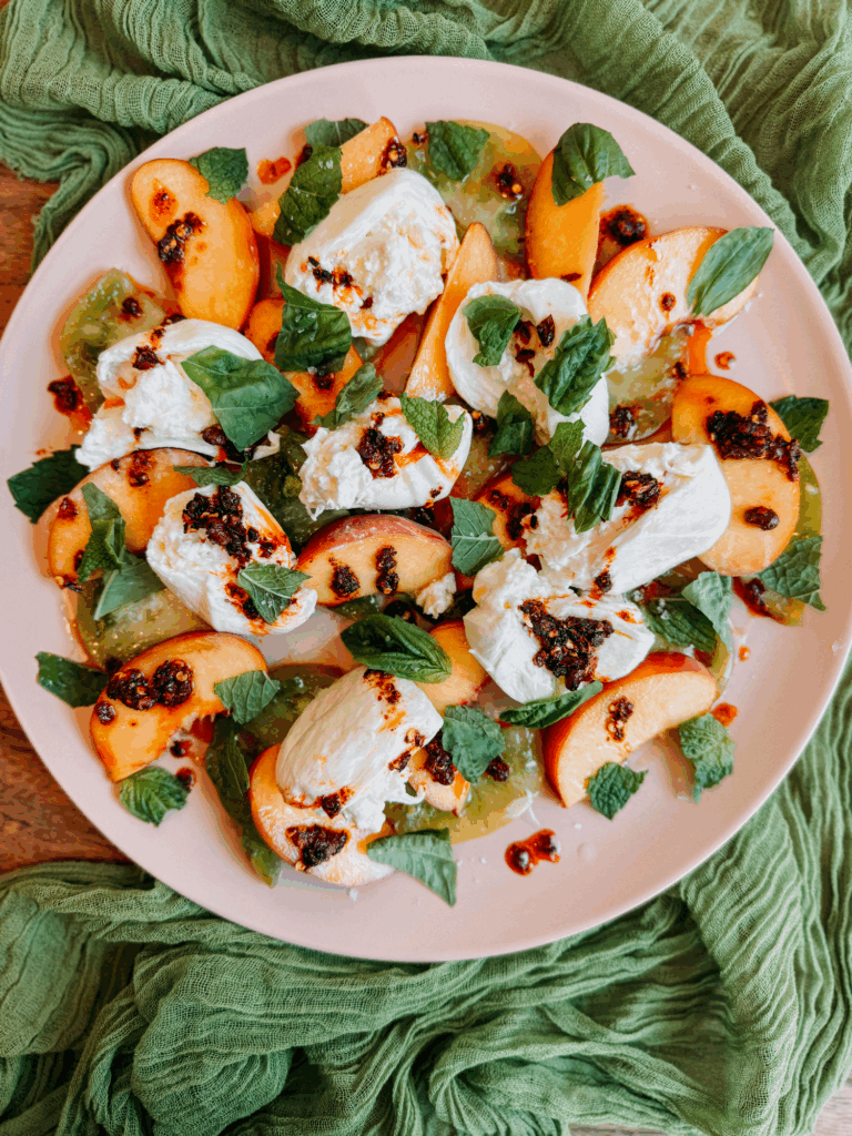 Overhead wide shot of burrata with peaches, tomatoes, chili crisp, and fresh herbs on a pink plate with green fabric.
