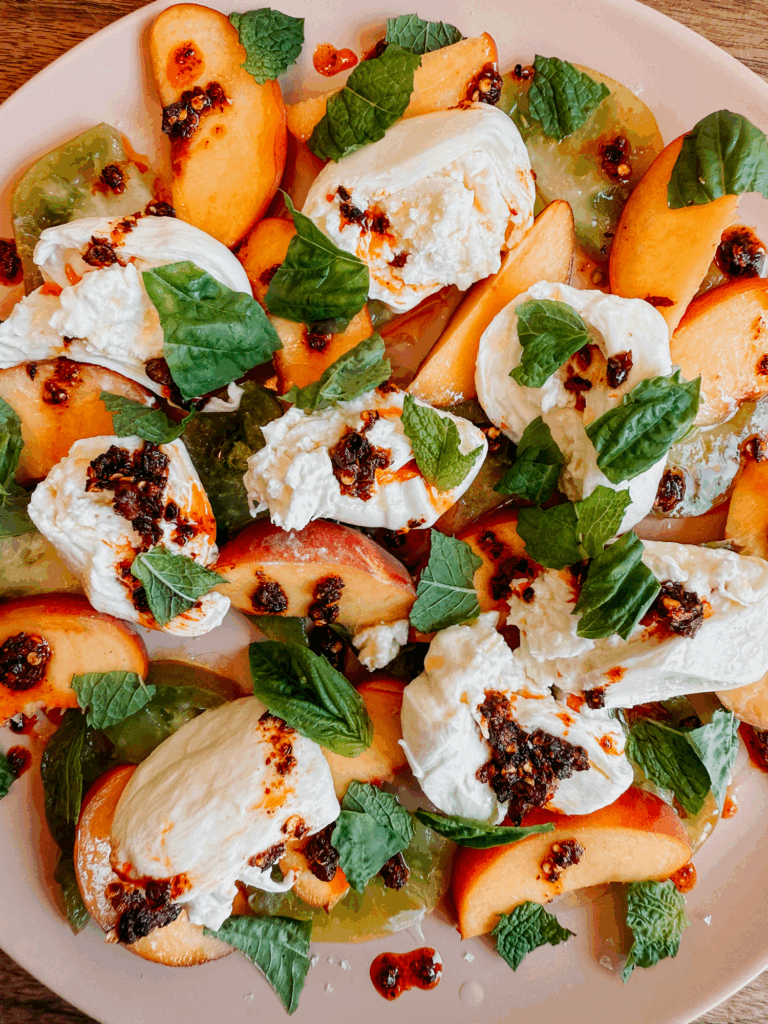 Overhead shot of burrata, peaches, tomatoes, and chili crisp arranged on a plate with fresh mint and basil.