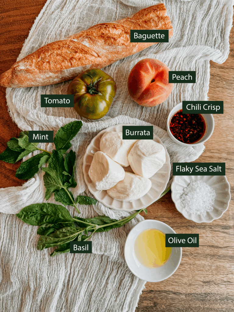Flat lay of ingredients for burrata with peaches, tomatoes, and chili crisp, including baguette, peach, tomato, burrata, basil, mint, chili crisp, olive oil, and flaky salt.