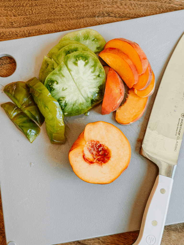 Sliced peaches and green heirloom tomatoes on a cutting board with a knife.