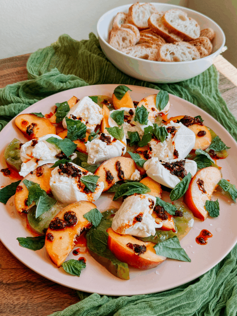 Platter of burrata with peaches, tomatoes, chili crisp, and herbs, served with a bowl of sliced baguette.