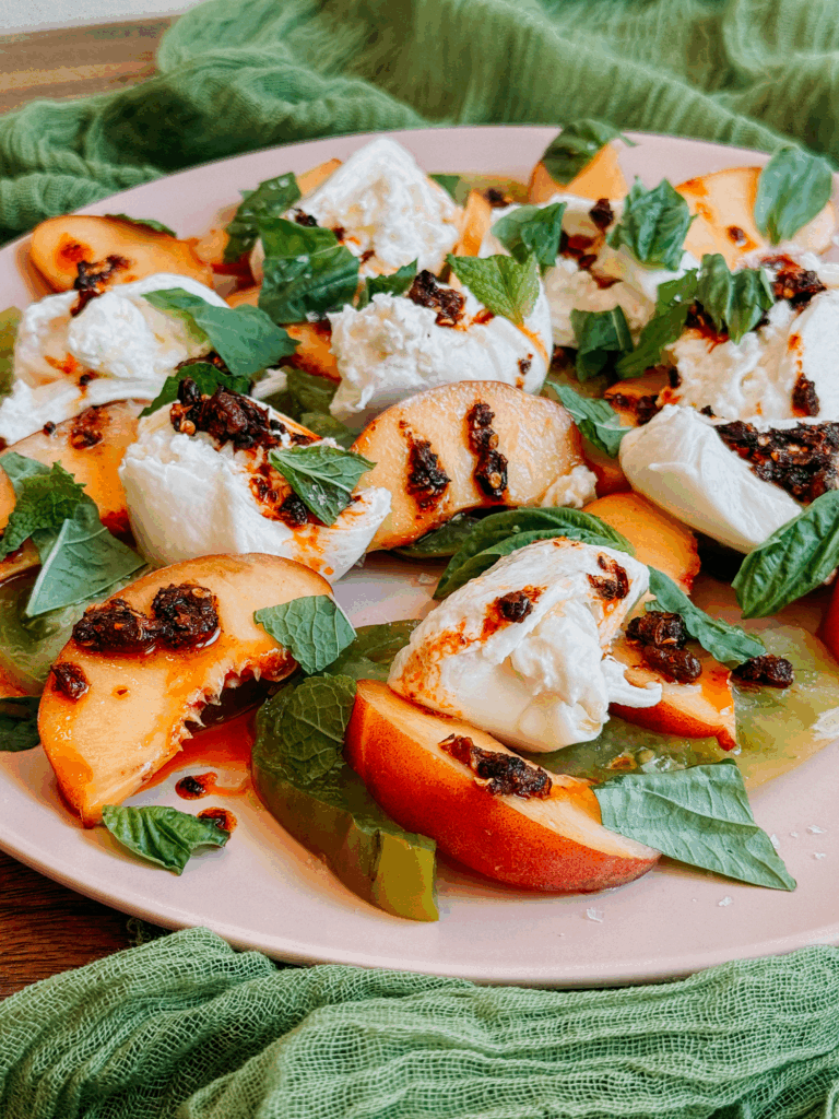 Close-up of burrata, peaches, tomatoes, and chili crisp garnished with basil and mint.