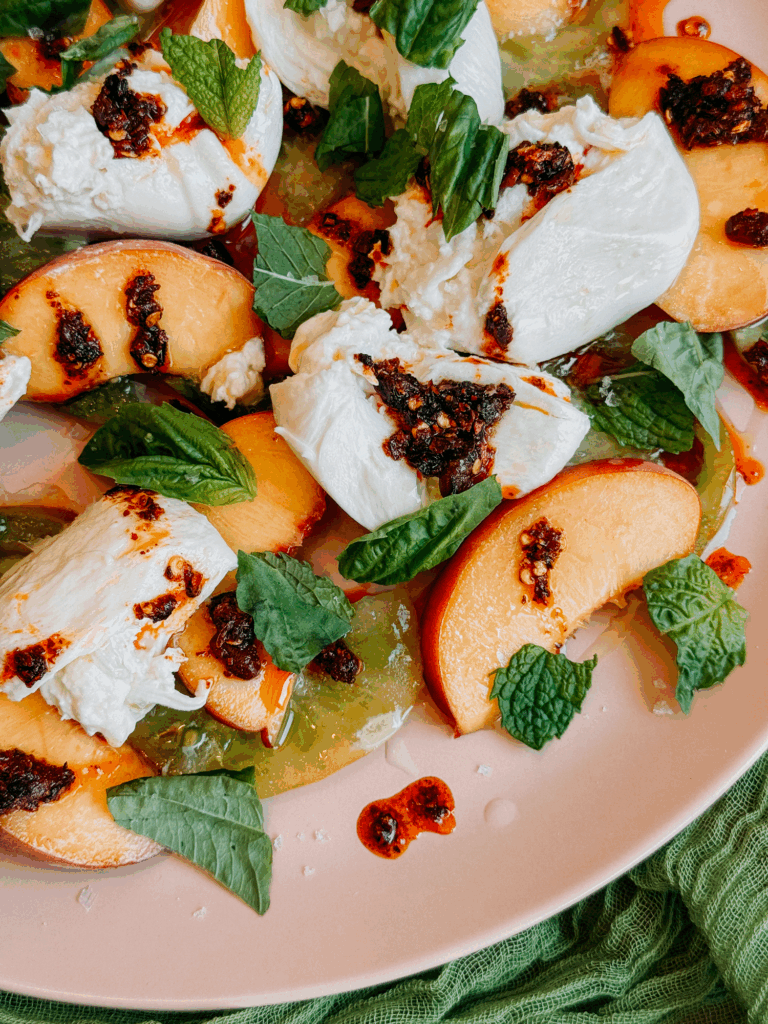Overhead close crop of burrata with peaches, tomatoes, chili crisp, and herbs on a pink plate.