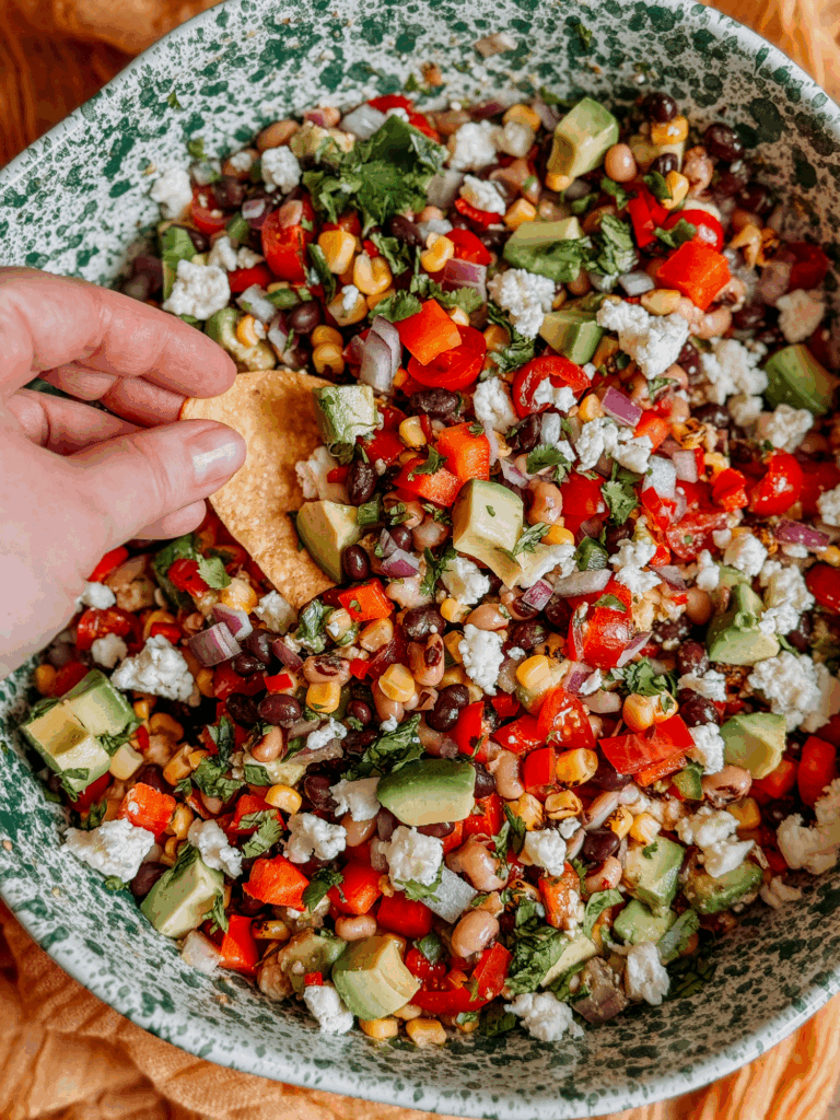 Hand dipping tortilla chip into bowl of Cowboy Caviar with avocado, corn, beans, and cotija cheese.
