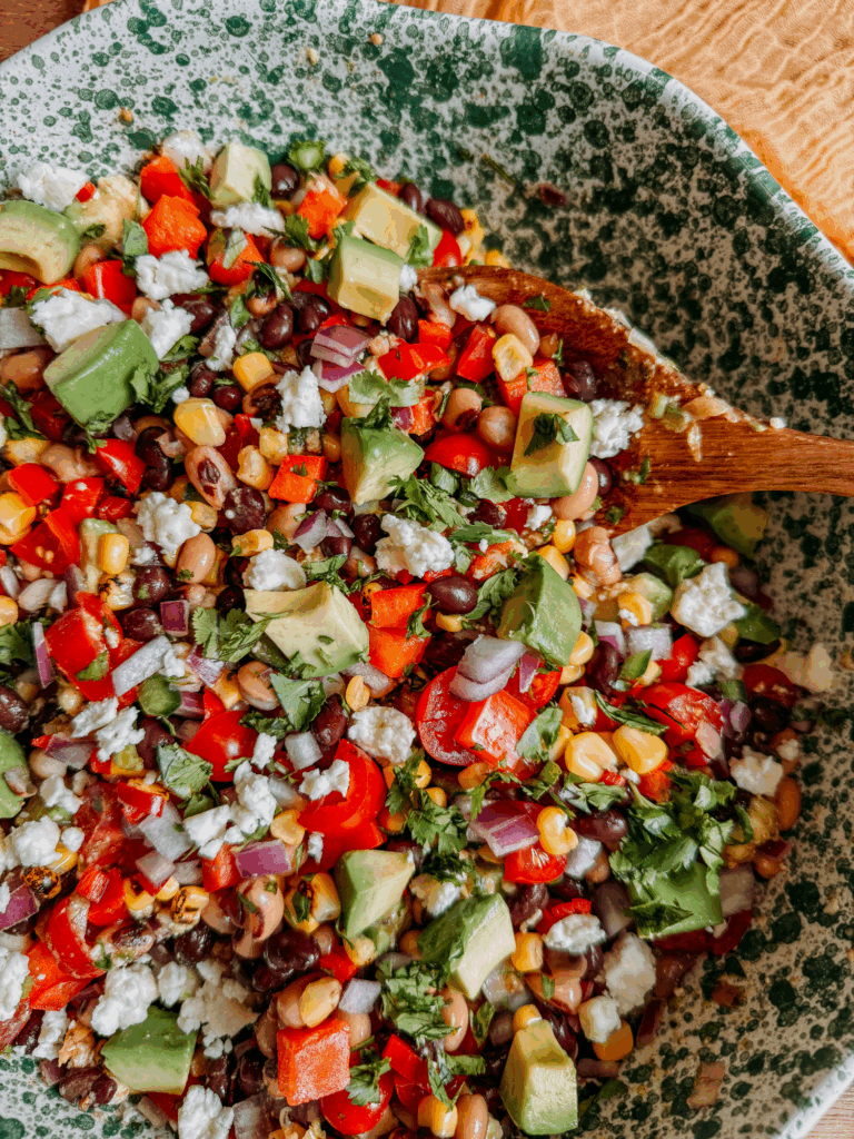 Cowboy Caviar bean salad with avocado, corn, black beans, black-eyed peas, peppers, onion, and cotija cheese in a green speckled bowl with a wooden spoon.