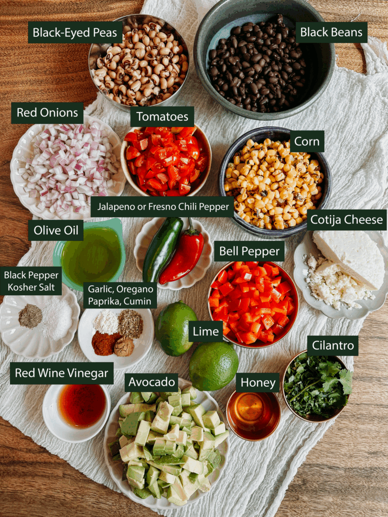 Ingredients for Cowboy Caviar laid out on table, including beans, corn, avocado, peppers, tomatoes, onion, lime, cilantro, and cotija.