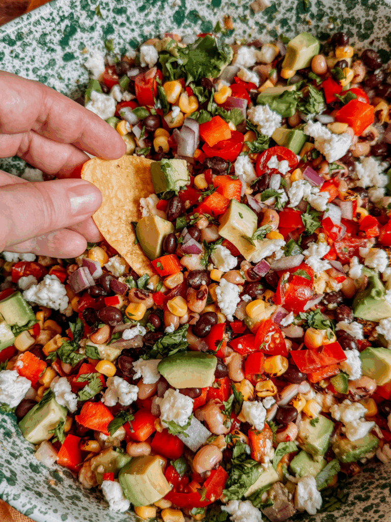 Hand scooping Cowboy Caviar with tortilla chip from bowl filled with avocado, beans, corn, peppers, and cotija cheese.