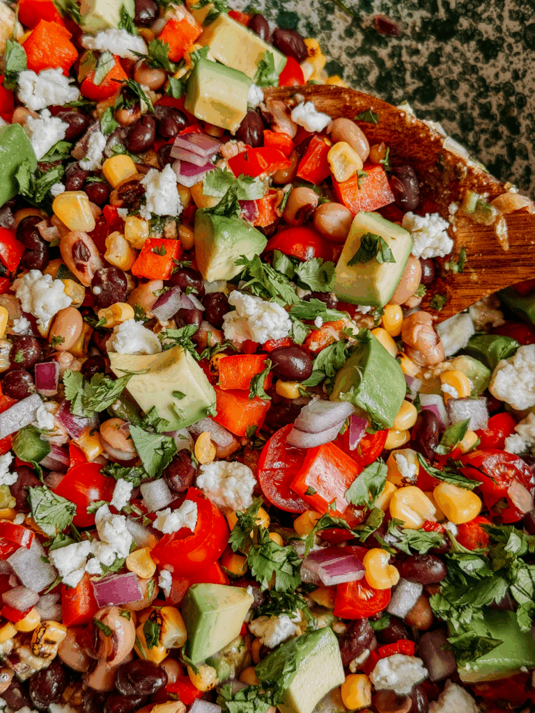 Wooden spoon in bowl of Cowboy Caviar with avocado, corn, black beans, black-eyed peas, tomatoes, peppers, and cotija cheese.